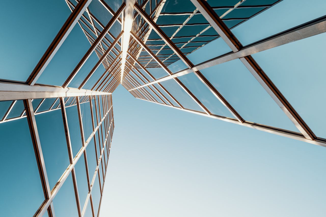 From below of multistory geometric building with glass mirrored windows reflecting cloudless blue sky on sunny day